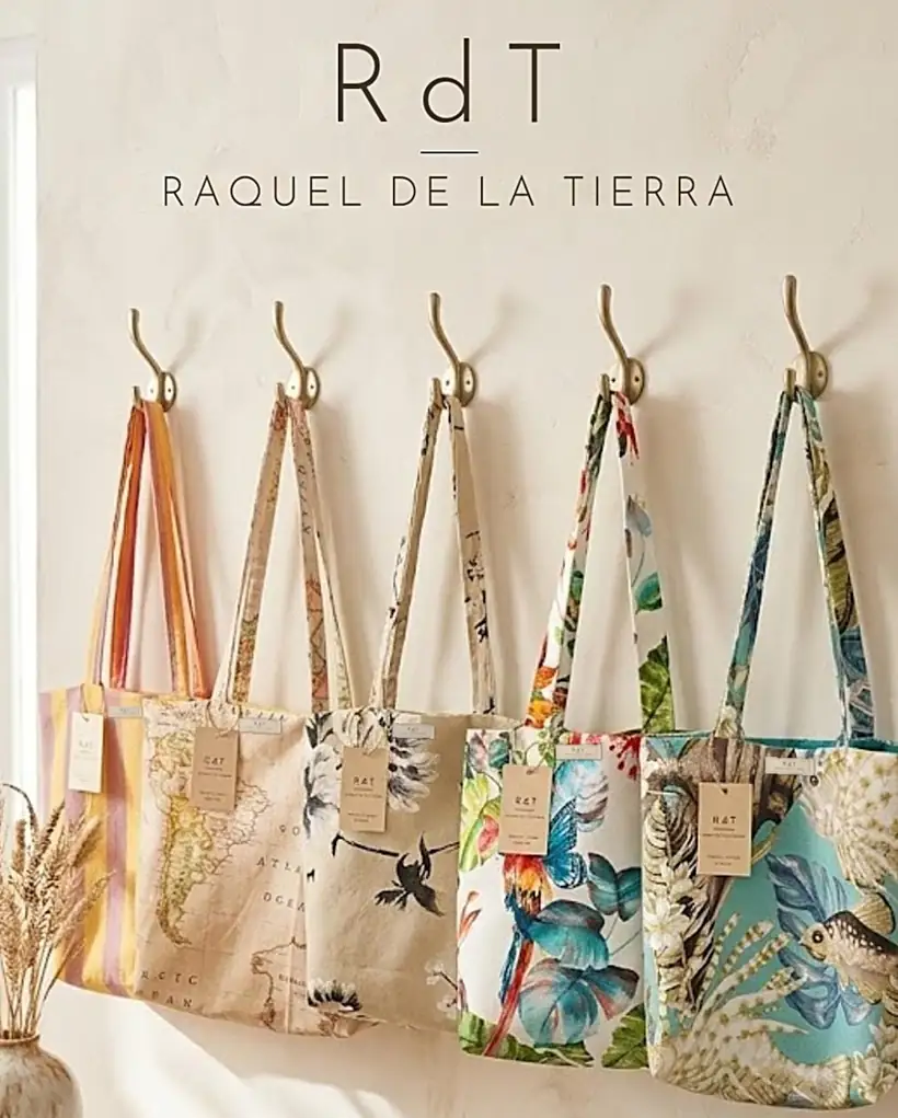 Cinco totes de Raquel de la Tierra colgadas de ganchos dorados: rayas, mapamundi, flores en negro, guacamayo y fondo marino